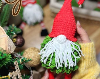 May include: A handmade crochet gnome with a red pointed hat, white beard, green body, and red legs. The image includes the text "Gnomes" and "Pattern by TwoBoyCrochet PDF English". The gnome is held in a person's hands.