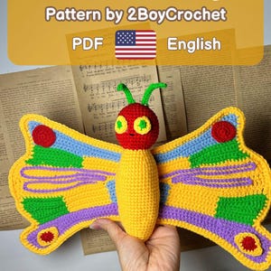 May include: A colorful crochet butterfly with yellow, blue, green, and purple wings. The butterfly has a red head and a yellow body. The image includes the text "Butterfly" and "Pattern by 2BoyCrochet PDF English".