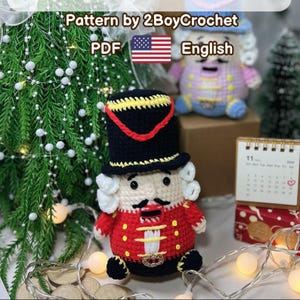 May include: A handmade crocheted nutcracker doll in red, black, and yellow, with a black hat and white hair. The doll is in front of a Christmas tree. The text "Pattern by 2BoyCrochet PDF English" is visible.