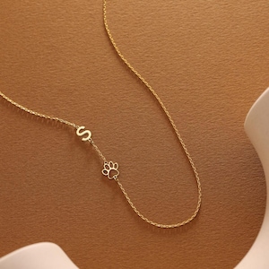 Puede incluir: Un delicado collar de oro con un dije de huella de pata y la letra "S". El colgante cuelga de una fina cadena, creando un diseño minimalista y elegante. El collar se exhibe sobre un fondo cálido y neutro.
