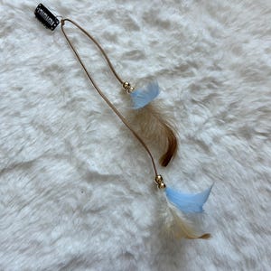 Può includere: Un accessorio per capelli con una clip nera, un cordino marrone e piume decorative. Le piume sono marroni chiare e azzurre, con perline dorate. Progettato per essere indossato nei capelli.