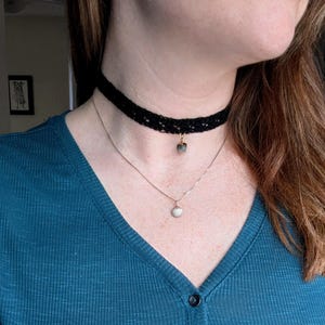May include: A black lace choker necklace with a small pendant, and a delicate silver chain necklace with a white pendant. The person is wearing a teal blue long-sleeved top.