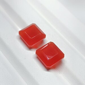 May include: Two square-cut, vibrant red gemstones with faceted edges. The gems are displayed on a white, textured surface, showcasing their polished appearance and rich color. These could be used for jewelry making.