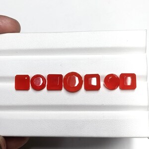 May include: A collection of seven polished, red gemstones in various shapes: square, round, and rectangular. The gemstones are displayed on a white, ridged surface, showcasing their vibrant color and cut.