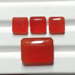 May include: Four polished, rectangular, red gemstones are displayed on a white surface. The gemstones have a smooth, reflective surface and are arranged in a rectangular pattern. The largest stone is at the bottom.