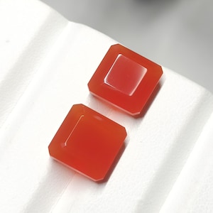 May include: Two square, orange-red gemstones with beveled edges are displayed. One gemstone has a square cut-out in the center. The gems are set against a white, textured surface, creating a striking contrast.