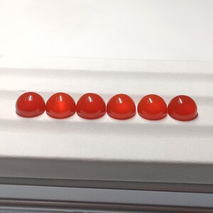 May include: Six polished, cabochon-cut gemstones in a vibrant, translucent red hue. The stones are arranged in a row, each with a smooth, rounded top and a flat base. The gemstones are displayed on a white surface.