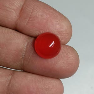 May include: A vibrant, polished, round, red gemstone. The gemstone is a solid, opaque color with a smooth, reflective surface. The gem is held in a hand, with the background out of focus.