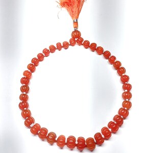 May include: A necklace featuring carved orange beads with a ribbed design. The beads are strung together with white string. The necklace has an orange tassel with a silver-colored metal accent at the top. The beads are approximately 0.5 inches in diameter.
