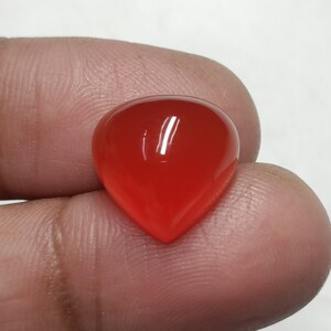 May include: A polished, teardrop-shaped gemstone in a vibrant red hue. The gem has a smooth, reflective surface, catching the light. The stone is held between fingers, showcasing its size and shape. This gemstone could be used for jewelry or other crafts.