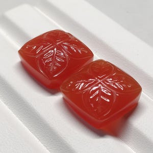 May include: Two square, reddish-orange gemstones with leaf carvings. The gemstones are set on a white surface. The leaf design is carved into the surface of each gemstone, creating a detailed and textured appearance. The gemstones are likely for jewelry making.