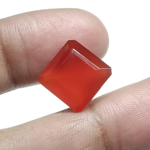 May include: A faceted, square-cut, red gemstone. The gem is held between fingers, showcasing its polished surfaces and vibrant color. The gemstone's shape is a square with beveled edges, reflecting light.