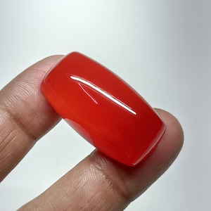 May include: A vibrant, rectangular, polished, red gemstone. The gem is held between two fingers against a white background. The stone has a glossy, reflective surface.