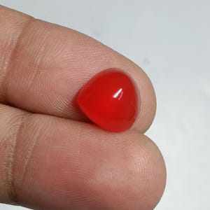 May include: A vibrant, polished, red gemstone with a smooth, rounded triangular shape. The gem is held between fingers against a plain white background, highlighting its glossy surface and rich color. This gemstone is suitable for jewelry making.