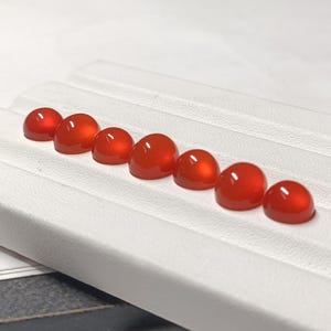 May include: Seven polished, cabochon-cut gemstones in a vibrant orange-red hue are arranged in a row. The stones are displayed on a white tray, showcasing their smooth, rounded surfaces and glossy finish. The gemstones are likely for jewelry making.