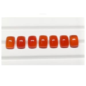 May include: Seven polished, rectangular orange gemstones are arranged in a row. The stones have rounded edges and a glossy finish, reflecting light. The gemstones are displayed on a white surface.