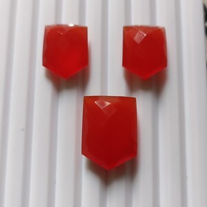 May include: Three faceted, shield-shaped, red gemstones are arranged on a white, ridged surface. The gemstones have a vibrant, translucent appearance, catching the light with their multiple facets. The overall composition suggests jewelry components or decorative elements.