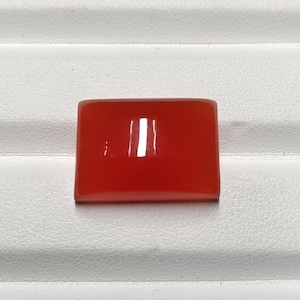May include: A rectangular, polished, red gemstone with a glossy finish. The gemstone is a solid, vibrant color and appears to be a cabochon cut. The stone is set against a white, textured surface.