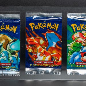 Puede incluir: Tres paquetes sellados de cartas coleccionables Pokémon exhibidos en un soporte acrílico transparente. Cada paquete presenta una ilustración de personaje Pokémon diferente sobre un fondo azul, con el logotipo de Pokémon en amarillo y verde. Los paquetes están etiquetados como "Trading Card Game" y "11 cartas canjeables".