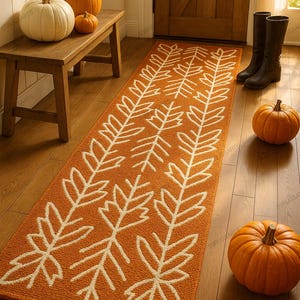 May include: An orange runner rug with a white leaf pattern. The rug is placed in a hallway with pumpkins and a wooden bench. The scene evokes a fall or autumn theme, with a warm and inviting atmosphere.