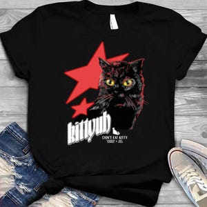 Könnte beinhalten: Schwarzes T-Shirt mit einer Grafik einer schwarzen Katze mit gelben Augen und einem roten Stern. Der Text "kittyub" steht über der Katze, darunter "DON'T EAT KITTY" und "CASEOH EST. 2022". Das Shirt wird auf einer Holzoberfläche präsentiert.