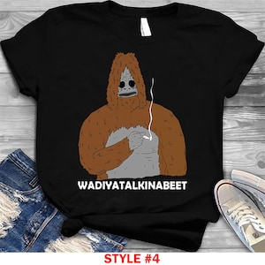 Peut inclure: T-shirt noir avec une illustration de dessin animé d'une créature ressemblant à Bigfoot en train de fumer. La créature est brune avec un visage et une poitrine gris. Le texte "WADIYATALKINABEET" est imprimé en dessous. Le texte "STYLE #4" est imprimé en bas.