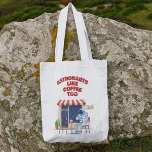 Op de afbeelding: Witte draagtas met de rode tekst "ASTRONAUTS LIKE COFFEE TOO". De afbeelding toont een astronaut in een café. De tas heeft twee witte handvatten en is gemaakt van stevig materiaal.