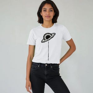 Pode incluir: T-shirt branco com um gráfico preto e branco de um planeta e o texto "INTERSTELLARWEAR" impresso verticalmente. A modelo está vestida com jeans pretos. Uma peça de roupa casual.