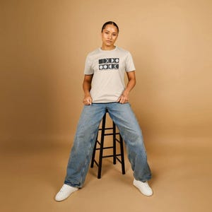 以下が含まれることがあります： 長方形と円の黒いグラフィックデザインが施された、明るいベージュのTシャツ。青いジーンズと白いスニーカーを履いた人が、黒いスツールに座っています。背景は無地のタン色です。