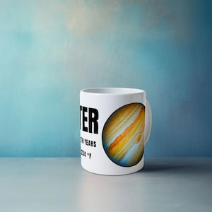 Può includere: Tazza in ceramica bianca con un'illustrazione colorata di Giove e la parola "JUPITER" in grassetto nero. Il testo aggiuntivo include "12 EARTH YEARS" e "234 °F". La tazza ha un manico bianco ed è ambientata su uno sfondo blu.