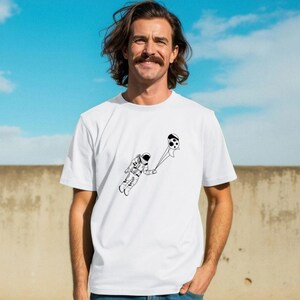 May include: White t-shirt with a black and white astronaut and football graphic. The illustration shows the astronaut floating with the ball. The tee is made of a soft material and is suitable for casual wear. It has a crew neck.