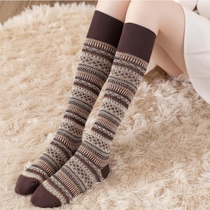 Può includere: Calze al ginocchio con polsino, tallone e punta marroni. Le calze presentano un motivo geometrico nei toni del marrone, beige e crema. Le calze sono esposte su un soffice tappeto color crema.