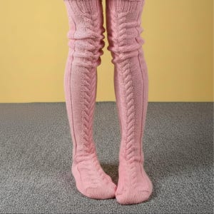 May include: A pair of light pink over-the-knee socks featuring a vertical cable knit pattern running down the center of each leg. The socks are constructed from a textured knit fabric with ribbed cuffs at the top and a reinforced heel and toe area.