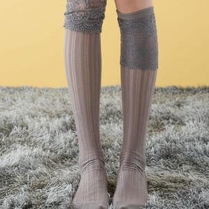 May include: A pair of knee-high socks in a muted gray tone featuring a vertical ribbed texture throughout the leg. The upper portion of each sock is finished with a wide, decorative band of intricate gray lace trim that sits below the knee.