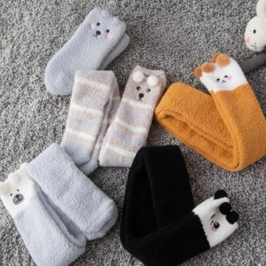 May include: A collection of fuzzy, plush socks featuring various animal designs. The set includes grey, striped, mustard yellow, and black socks, each adorned with embroidered faces, small ears, or pom-poms at the top cuffs for a soft texture.