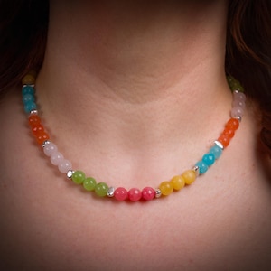 Könnte beinhalten: Eine Perlenkette bestehend aus einer Abfolge von mehrfarbigen, facettierten und glatten runden Edelsteinperlen in Rosa, Gelb, Orange, Blau und Grün, die durchgehend mit kleinen, polierten silberfarbenen Metallzwischenperlen verziert ist.