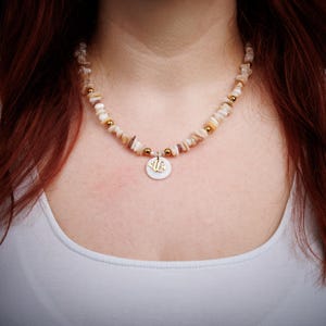 Könnte beinhalten: Eine Halskette mit einer Kette aus kleinen, unregelmäßig geformten, hellfarbigen Muschelkugeln. Die Halskette hat goldfarbene Akzente und einen runden Anhänger mit Fischmuster. Die Halskette wird über einem weißen Oberteil getragen.