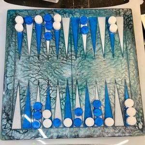 Puede incluir: Un tablero de backgammon con un diseño veteado en azul verdoso y blanco. El tablero presenta fichas de juego azules y blancas. Los triángulos son blancos y azules. El tablero es rectangular y tiene un acabado brillante.