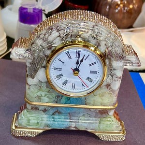 May include: A decorative clock with a gold-colored frame and a white clock face with black Roman numerals. The clock is embellished with rhinestones and features a clear resin design with embedded flowers and barber tools.