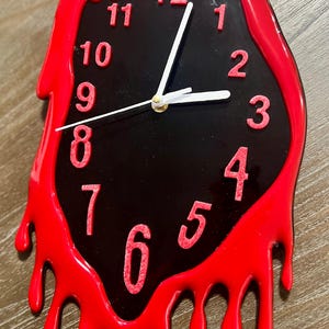 May include: A novelty wall clock with a black face and red, dripping frame. The clock features white hands and red numerals. The design gives the appearance of melting or dripping paint, creating a unique, eye-catching aesthetic.