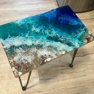 May include: A rectangular side table with a resin ocean design. The tabletop features a blue and turquoise wave pattern with white foam, seashells, and starfish. The table has dark metal hairpin legs.