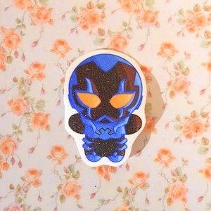 Könnte beinhalten: Ein Aufkleber mit einer stilisierten Chibi-Figur in einem blau-schwarzen Rüstungsanzug, markanten orangefarbenen Augen und einer glitzernden Textur. Die Figur hat einen abgerundeten Kopf und ein kompaktes Design mit deutlichen blau-schwarzen Segmenten.
