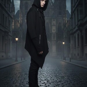 K&ouml;nnte beinhalten: Eine Person in einem schwarzen Kapuzenmantel und einer schwarzen Hose steht auf einer Kopfsteinpflasterstra&szlig;e. Der Mantel hat ein asymmetrisches Design mit Rei&szlig;verschluss. Im Hintergrund eine gotische Kathedrale und Stra&szlig;enlaternen.