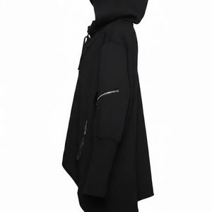 K&ouml;nnte beinhalten: Schwarzer Kapuzenponcho mit einzigartigem asymmetrischem Design. Mit gro&szlig;er Kapuze, langen &Auml;rmeln mit Rei&szlig;verschlussakzenten und einer drapierten, flie&szlig;enden Silhouette. Ein modernes, kantiges Oberbekleidungsst&uuml;ck.