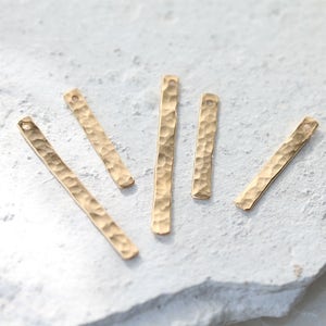 Peut inclure: Cinq pendentifs en métal de couleur dorée avec une texture martelée. Chaque barre a un trou en haut pour l'attacher à des bijoux.
