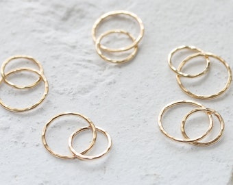 May include: Several gold-colored, hammered-texture, interlocking circle charms are displayed against a white, textured surface. The charms are arranged in pairs, with one smaller circle nested within a larger one. The overall theme is jewelry.