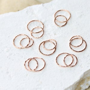 May include: A set of eight rose gold stacking rings. The rings are thin and delicate, with a hammered texture. They are arranged in a random pattern on a white surface.