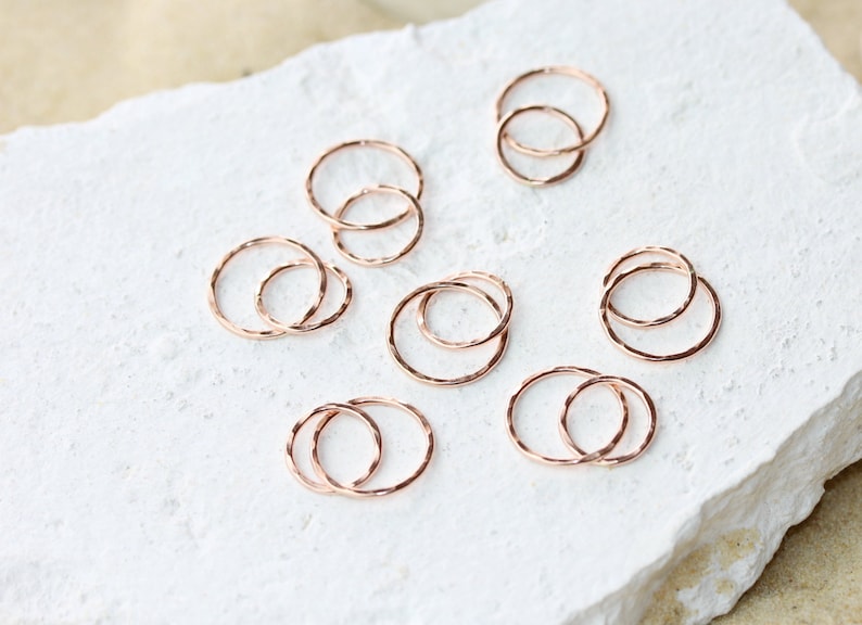 May include: A set of eight rose gold stacking rings. The rings are thin and delicate, with a hammered texture. They are arranged in a random pattern on a white surface.