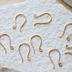 Könnte beinhalten: Nahaufnahme von acht goldfarbenen Ohrhaken auf einer weißen, strukturierten Oberfläche. Die Haken sind aus Metall und haben ein schlichtes, elegantes Design.