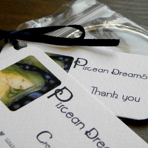 May include: Two white cards with the text "Piscean Dreams" and "Thank you" on them. The cards are on top of a clear plastic bag with a silver ring inside. The top card has a small illustration of a woman with blue and green hair.